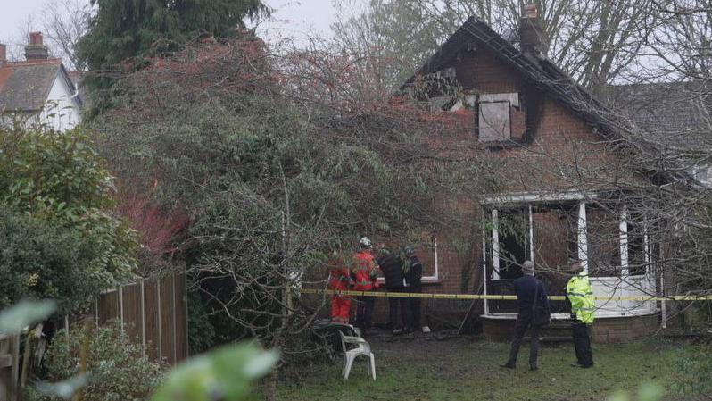 A house which has windows boarded up and appears damaged. In front of the house, there is overgrown trees. There is yellow tape across the front, with a police officer and a man standing in front of it. Behind the tape, four people can be seen, two wearing orange high vis jackets. In the foreground, there is grass and a garden chair, and a fence to the left of the image. 