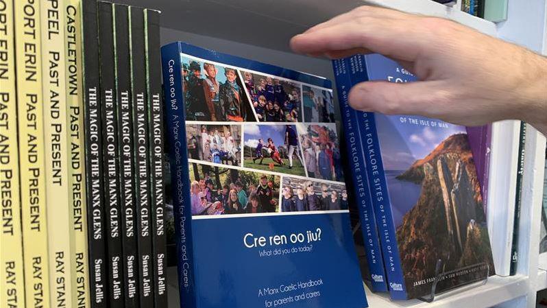 The Cre Ren Oo Jiu? book sits on a bookshelf. It is blue and has pictures on children doing activities, such sa playing sport. There are other books on folklore and glens that sit next to it.