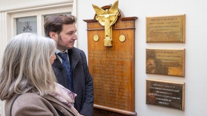 A woman with shoulder-length grey hair, wearing a lilac coat, and a man with short dark hair, wearing a blue tie and a dark coat, are looking at some brass plaques on a wall. There is also a wooden board titled: "Roll of Honour".