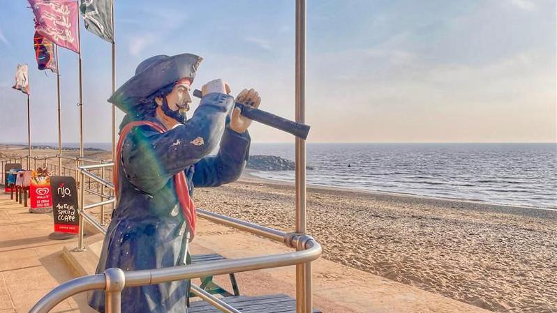 A pirate statute is standing next to a metal barrier. It has a telescope, and is looking out to the sea. 