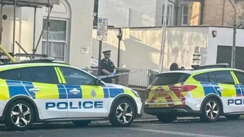 Two police cars with a police office standing behind a police cordon. The police officer, wearing black, stands in front of a cream coloured house.