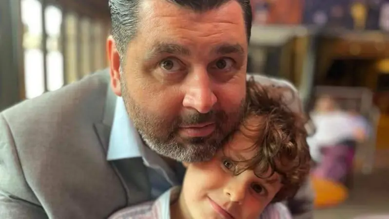 A man and a boy look directly in to the camera lens. They are smiling and both have brown eyes and brown hair. The background is out of focus. The man is wearing a suit.