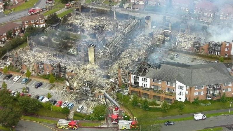 An aerial view showing the ruins of an apartment complex which has been destroyed by a fire. Smoke can be seen rising from the ruins and three fire engines are parked nearby.