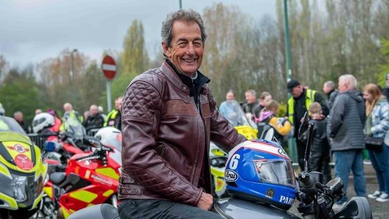 A man in a leather jacket sitting on a motorbike and smiling at the camera