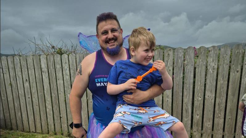 Thomas Davies dressed in purple with fancy dress fairy wings, smiling, holding a smiling Lyidan.