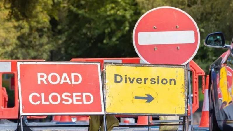 Road closure signs next to a road. There are two red signs and a yellow sign. There are also trees in the background.