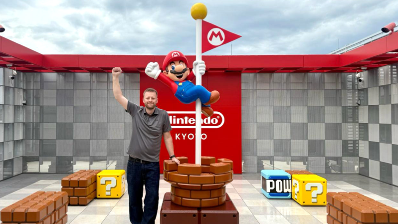 A man who is wearing a grey short-sleeved shirt and dark trousers is standing alongside a model of the computer game character Mario, with the words "Nintendo Kyoto" behind them. The man is raising his arm in the air, copying the pose of the model, who is wearing blue dungarees, a red top and a red hat.