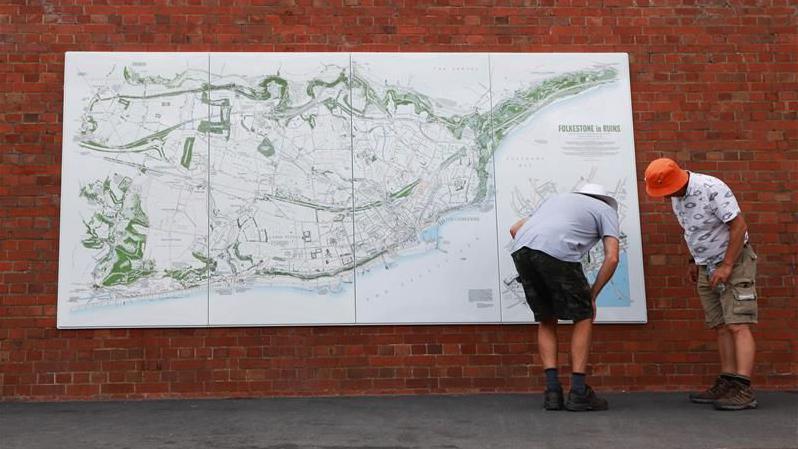 Two men in shorts look at a giant map on a wall in Folkestone
