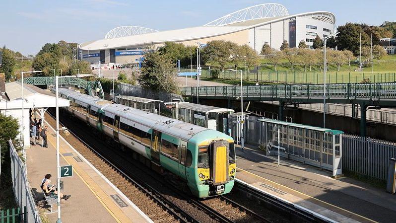 A Southern Rail train stopping at a station. There is a football stadium in the background, and a number of people waiting on the platform. 