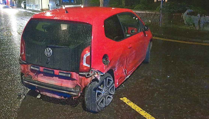 A red VW Up, which has been damaged at the back. It's rear driver-side tyre is deflated and the driver's side door is smashed in. Some of the bodywork at the back of the car has fallen off. 