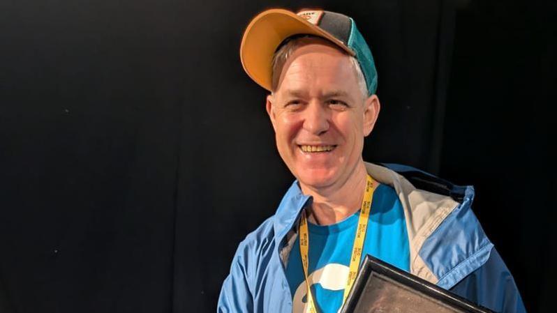 A man with grey hair, wearing a blue baseball cap, blue t-shirt and blue jacket, holding a black framed item, standing against a black background.