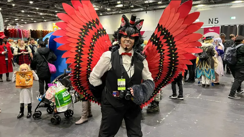 A fan wearing a costume comprised of a white shirt, a black waistcoat, large red wings and red facepaint.