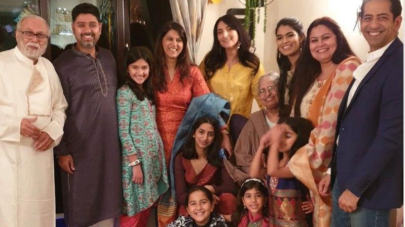 Eight members of the Bhatia family are standing up, there are three men and five women. At their feet are one woman and four children. They are all wearing bright outfits.