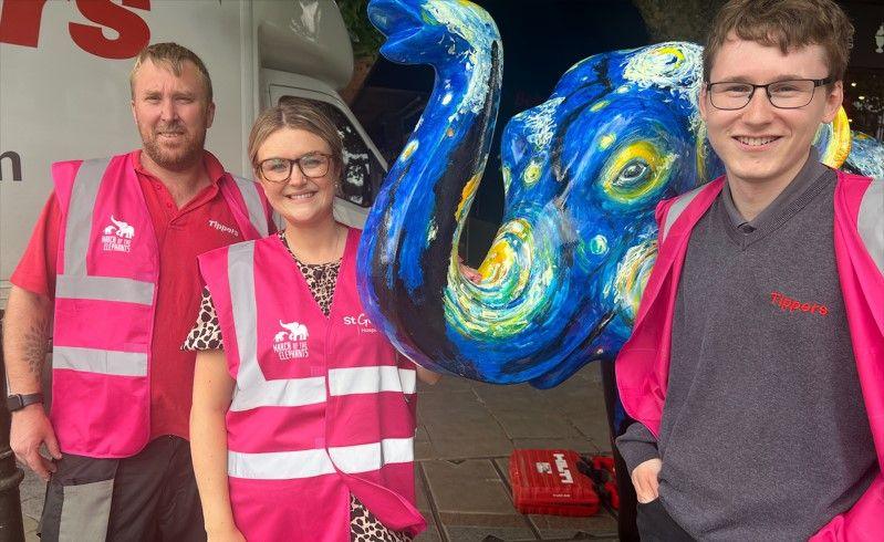 Two men and one woman in pink hi-vis St Giles jackets smile as they stand next to the Vincent Van Gogh themed elephant