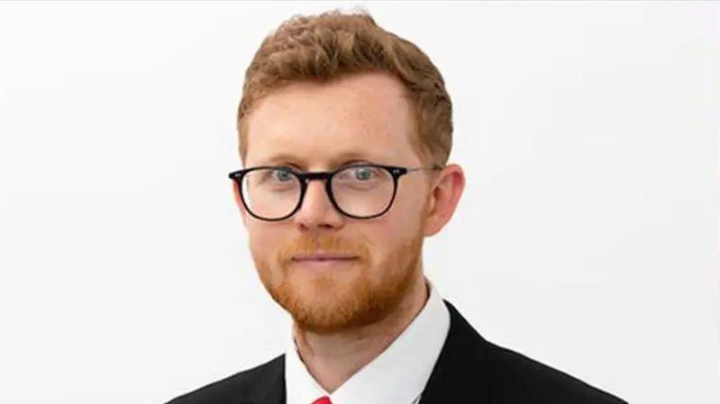 Headshot of Sam Gould in suit and red tie wearing glasses