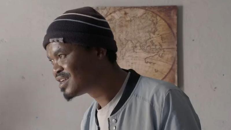 A side view of the head and shoulders of Sinqhiwe Goodman Sithole. He is wearing a dark beanie hat and grey jacket.