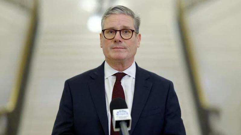 Sir Keir Starmer is wearing a navy suit with a red tie and white shirt. He is looking straight at the camera with a straight facial expression. He is wearing dark coloured glasses and has grey hair.