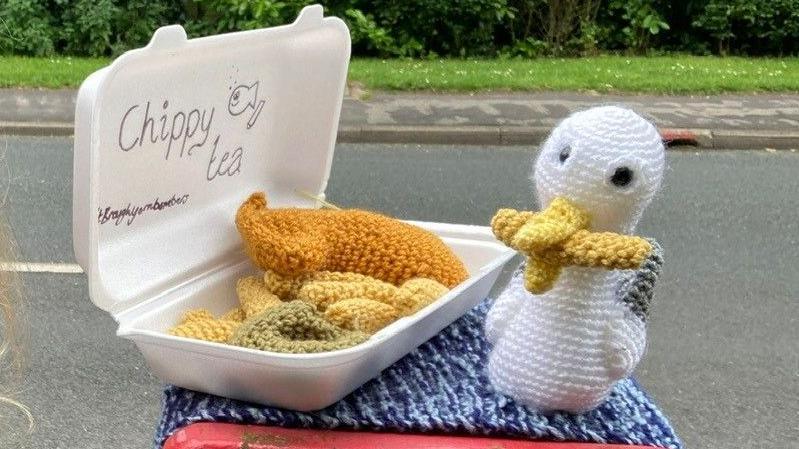 Knitted fish and chips are in a polystyrene food box.  A large knitted seagull sits to the side of it, with a large knitted chip in its beak.