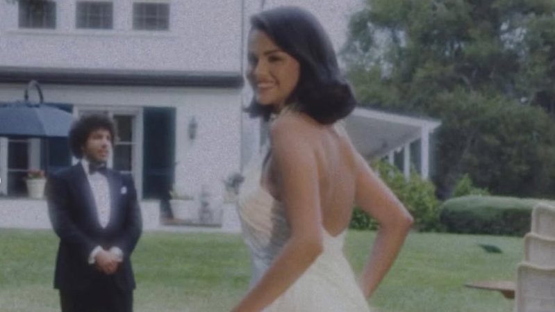 Selena Gomez, a young woman with dark hair, wears a sleeveless, high-necked white wedding gown and is smiling. In the background is Benny Blanco, a young man wearing a dark tuxedo and white shirt. They are standing in the grounds of a large white property.