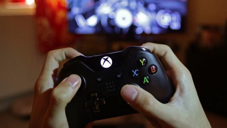 Two hands holding a black video game controller. The letters X, Y, B, A, are visible on the gadget. In the background is a blurred screen.