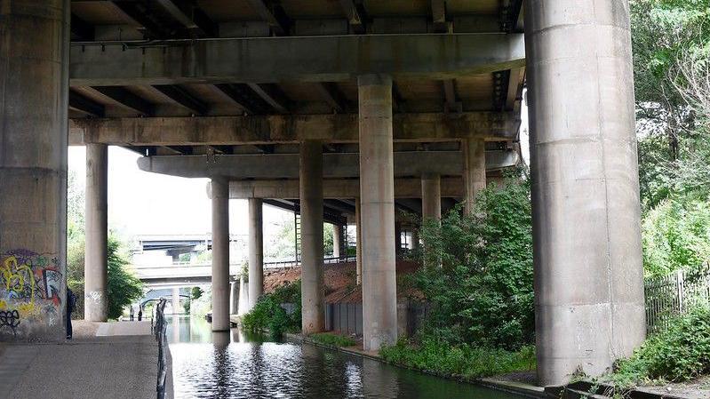 The huge beams underneath Spaghetti Junction. A canal flows underneath it and there is some graffiti. 