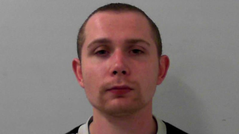 Mugshot of a man with a short goatee and a buzzcut. He has a neutral expression. He is wearing a grey jumper.