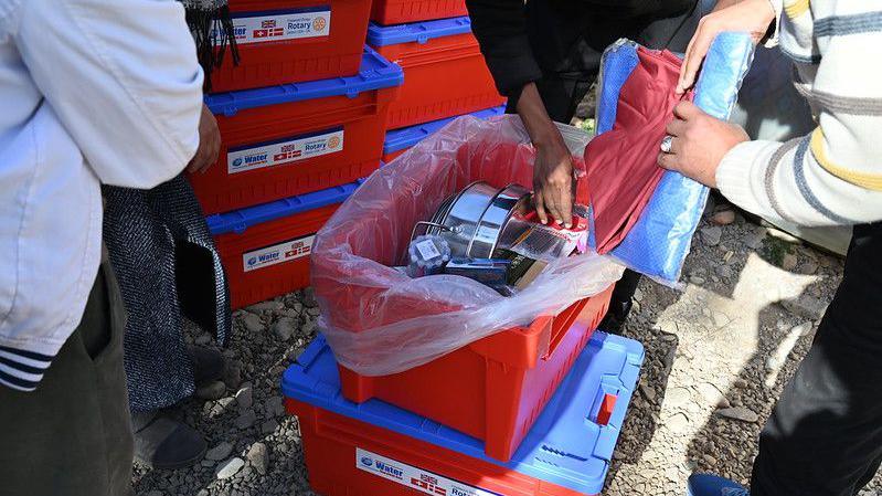 People looking through a red plastic box which contains pots and cooking utensils.