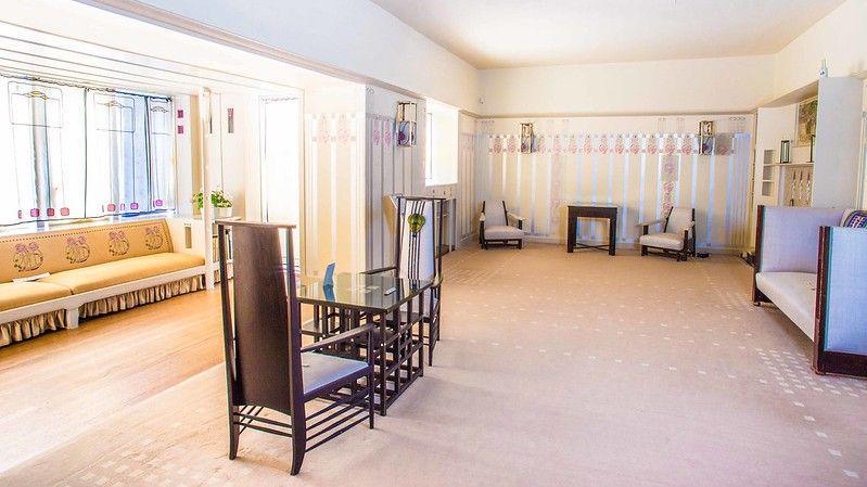 The inside of one of the rooms of the house. There is a traditional table and chairs in the middle with Mackintosh designs. There is a sofa and another traditional couch on both the left and right hand side of the picture. The walls are covered in a Mackintosh style wallpaper. 