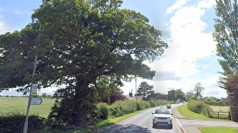 A Google Street View screenshot of the area where the care home and shops would be built. A country road is surrounded by fields and green open spaces. There are some houses in the distance and cars travelling towards the camera.
