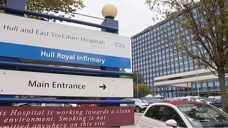 A blue and white sign directing visitors to the main entrance of Hull Royal Infirmary. The tall, glass-fronted hospital building is in the background. Cars are parked nearby.