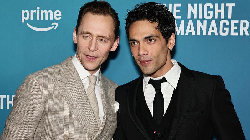 Tom Hiddleston stands on the left wearing a light suit. A man with dark hair and a dark jacket and tie stands to his left