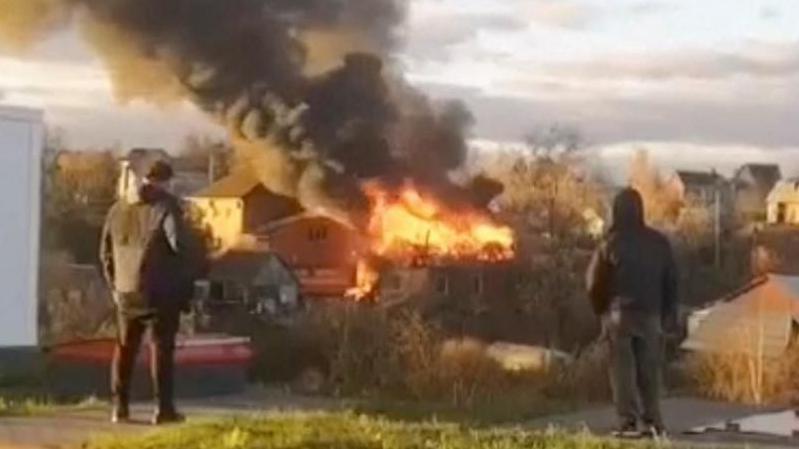 Grainy image of flames seen rising from debris in a residential area of Russia. Two people can be seen with their backs to the camera, looking at the scene of debris. Thick black smoke is rising from the flames.