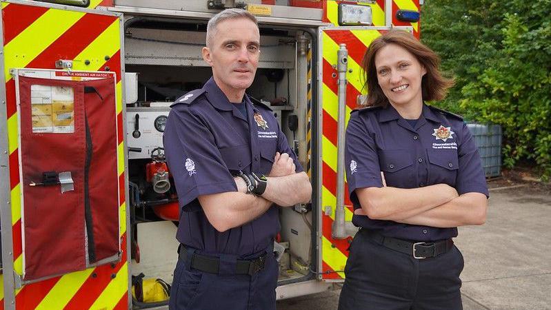 A male and female fighter wearing navy blue uniforms stand with their arms folded in front of the back of a fire engine