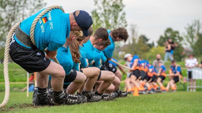 Teams of eight pull eight end of rope in a tug of war contest on a grass field. The competitor closest wears a German flag on the back of his blue polo shirt.