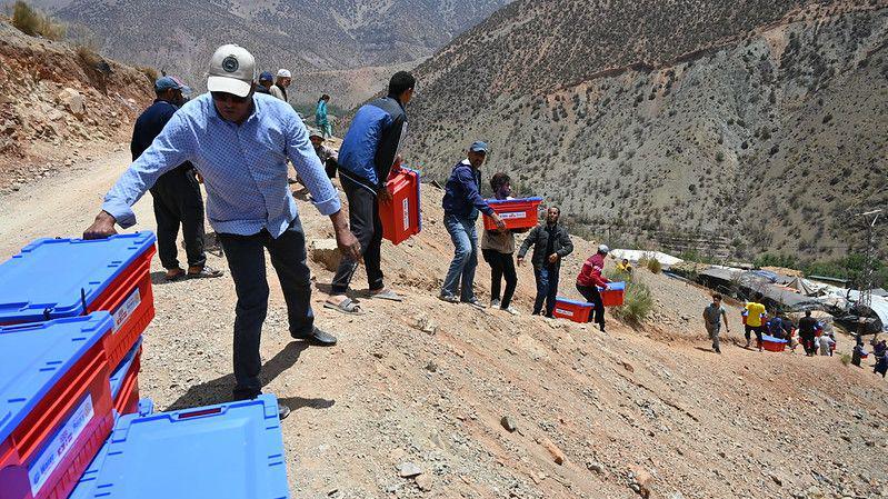 People carrying red and blue plastic boxes up a rocky mountain.