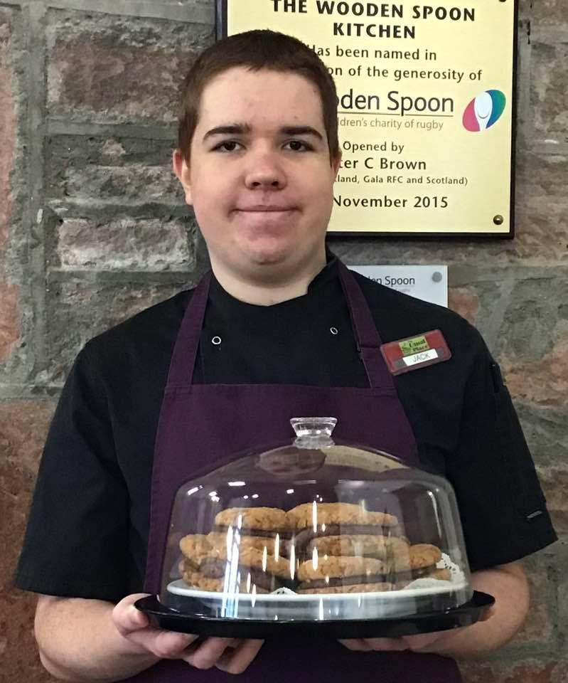 A young man with short dark hair in a black T-shirt and purple apron holds up a tray of cakes under a glass lid