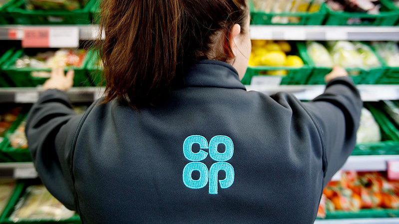 The back of woman with brown hair in a ponytail wearing a Co-op uniform with the logo on her back while reaching across shelves of vegetables