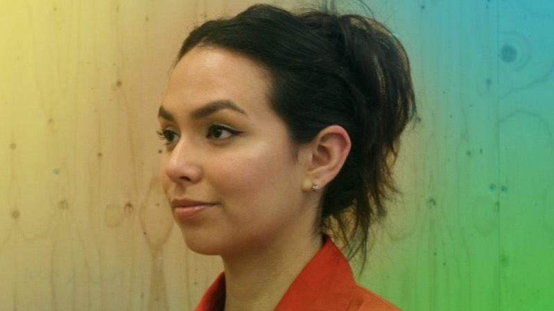 Edit Kruk, a woman wearing a red top, is standing against a wooden backdrop which is illuminated in light which fades from yellow to green and blue, left to right.