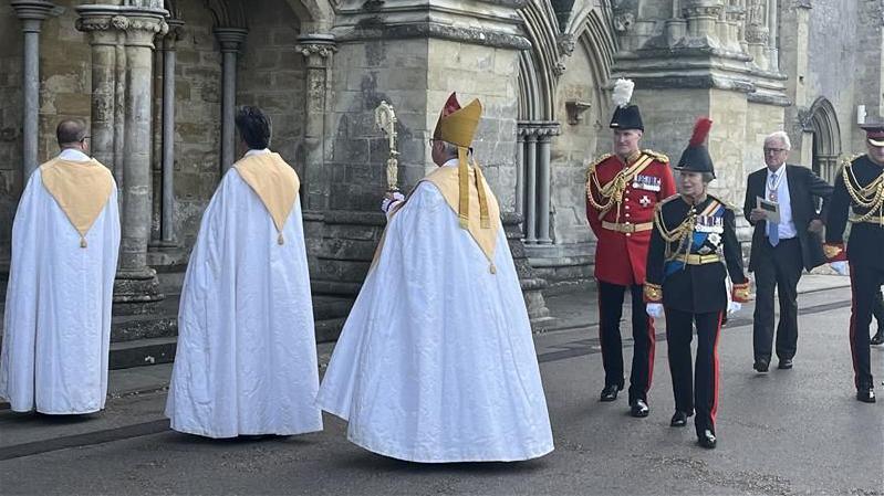 Princess Anne in black military dress and a hat with a red feather, entering Salisbury Cathedral flanked by three people in long white cloaks.