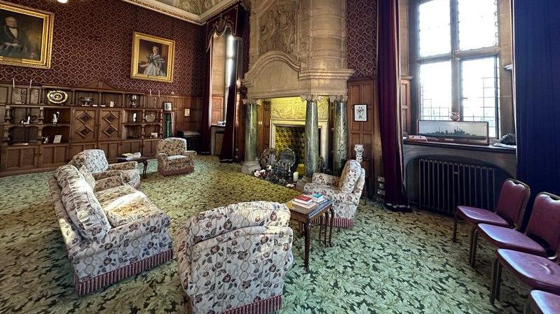 The Lord Mayor's Parlour is a grand room with dark wood-panelled walls, a carpet with a leaf design, very tall arched windows, a huge marble fireplace and an ornate ceiling. There is a selection of armchairs and a sofa around the fire place. On the walls are large antique paintings in gold frames of individual people and the ceiling has chandeliers.