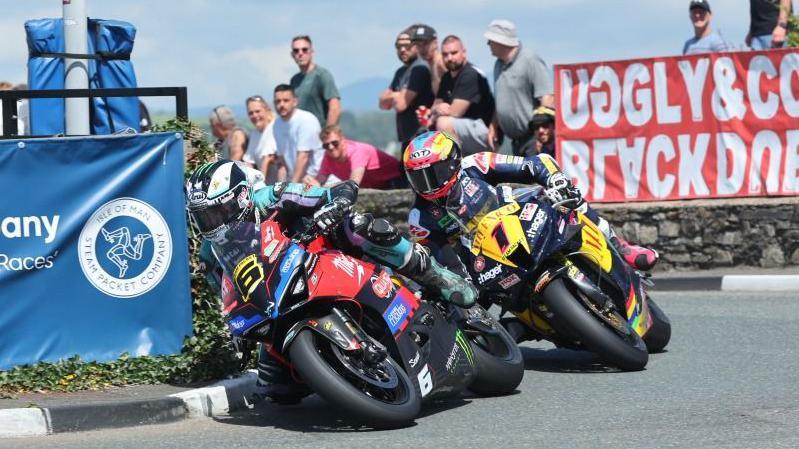 Michael Dunlop ahead of Davey Todd while rounding a corner past crash barriers as spectators watch on from behind.