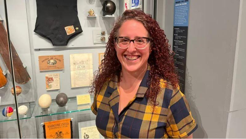 A woman with dark red long curly hair smiles at the camera. She is wearing a checked blue and yellow shirt and black glasses. She is standing in front of a museum shelving display
