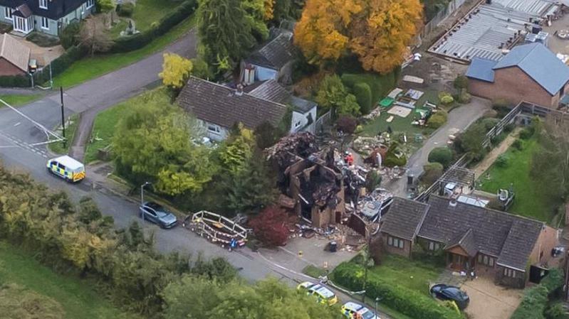 Drone photo shows destroyed buildings with emergency vehicles parked outside. Nearby homes appear undamaged.