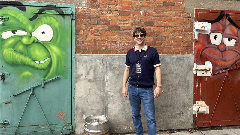 A man with short brown hair wearing black sunglasses, a navy polo shirt and blue jeans stands on a street. The wall behind him to his left and right have graffiti of green and red characters. 