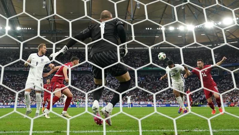 Action from England - Serbia Euro 2024 football match. England's Jude Bellingham heads a goal. Serbia's goalkeeper, wearing black, looks towards two England attackers and two Serbia defenders. Spectators are in the background.