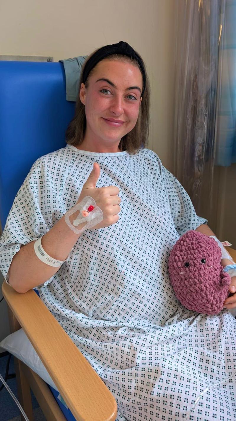 A woman with bobbed brown hair, sat in a hospital gown on a chair. She has a catheter in one hand, which she is holding up in a thumbs up. The other arm is cradling a crochet figure, in the shape of a pink kidney.