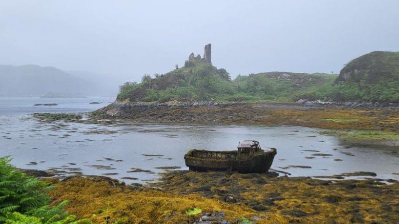 A boat rests up against rocks covered in kelp. In the background are castle ruins.