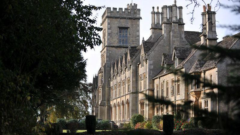 The buildings of the Royal Agricultural University bathed in autumn sunlight. The architecture of the building is Victorian gothic style. There are trees and bushes in the corners of the photograph.