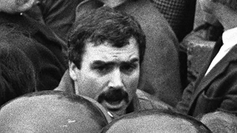 A black and white image of a man with dark hair and a moustache. 