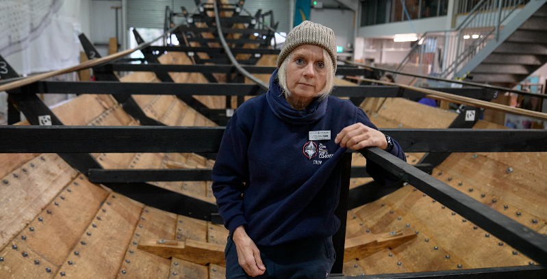 Jacq Barnard stands within the reconstructed ship's hull. One arm rests on black metal framework as she smiles at the camera. She is wearing a grey beanie hat, blue neck snood and a navy jumper with a name badge on her chest. 